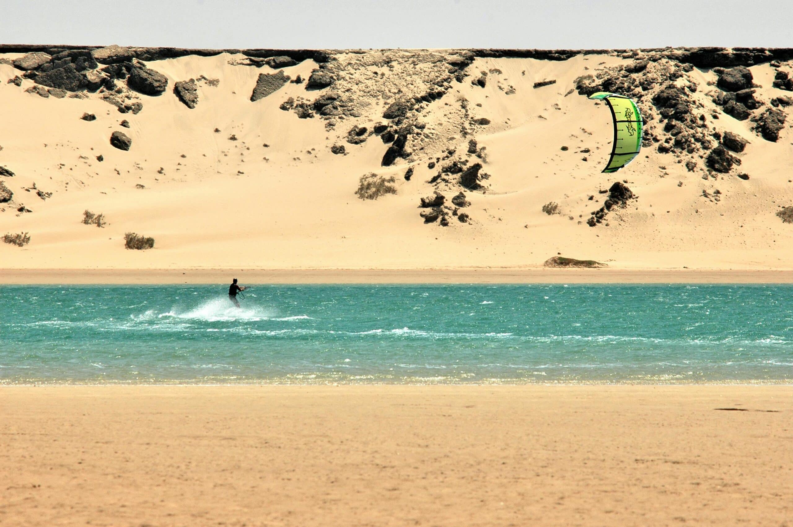 Une vue spectaculaire de Dakhla, où un kitesurfeur glisse sur les eaux turquoise, avec des dunes de sable désertique en arrière-plan.